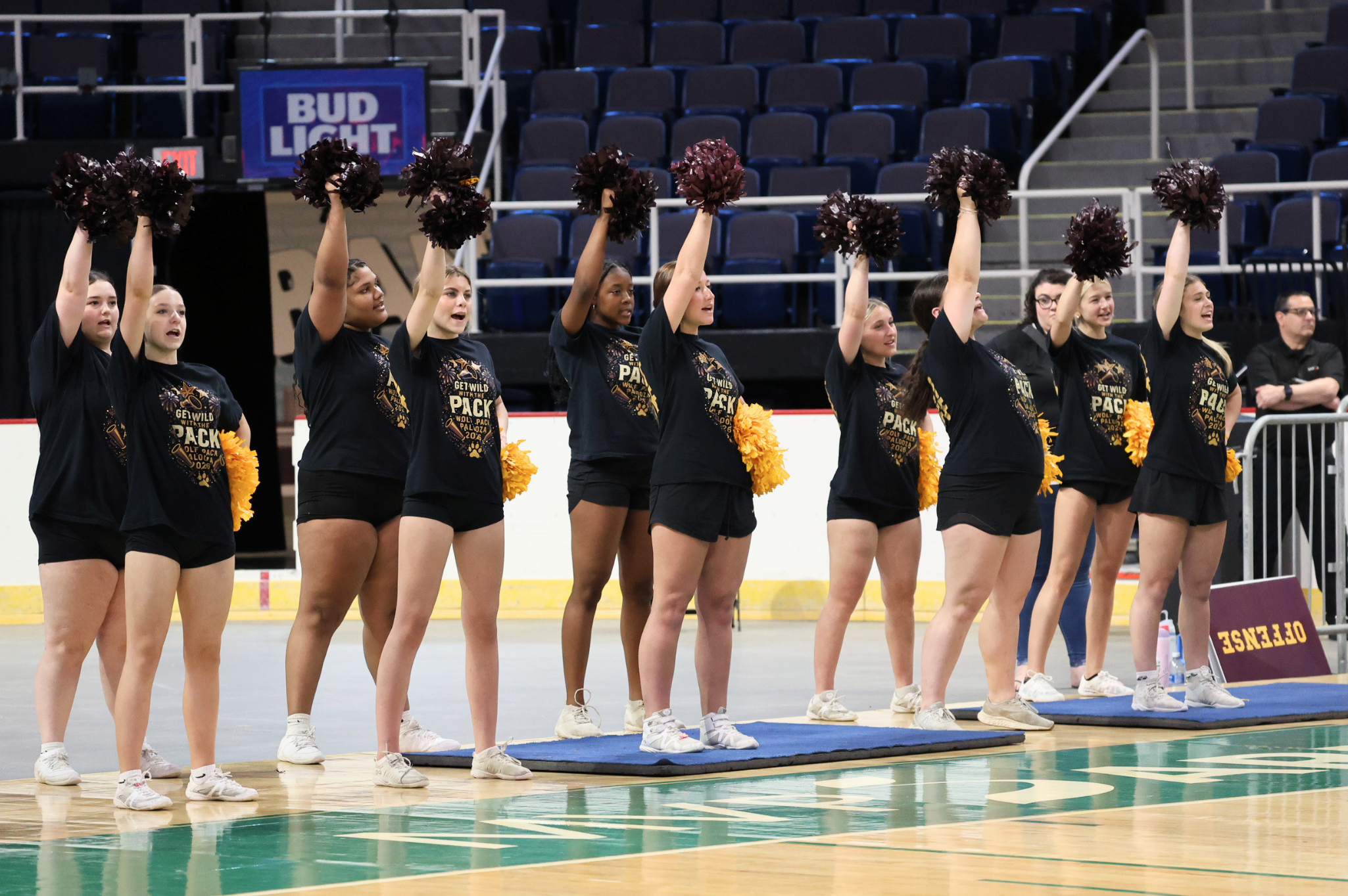 A line of cheerleaders wearing black shirts and holding up pom poms on the side of a basketball court